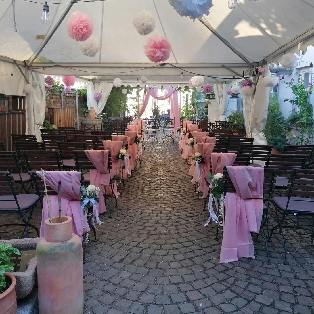 Hochzeitstrauung — Stuhlreihen mit rosa Stuhlhussen und weißen Pompoms unter Pergola